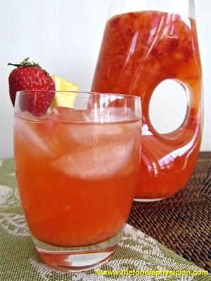 Glass of strawberry pineapple lemonade with a pitcher of it in the background.