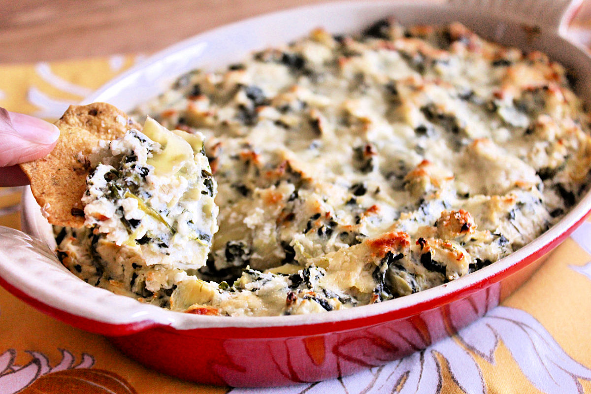 Taking a scoop of spinach artichoke dip out of a red casserole dish