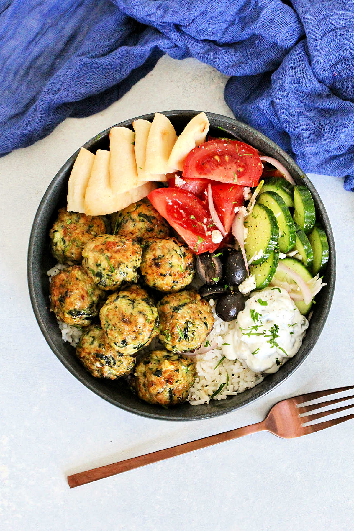 Overhead picture of Greek chicken meatballs in gray bowl with tzatziki, tomatoes, cucumbers, olives, feta and pita wedges.