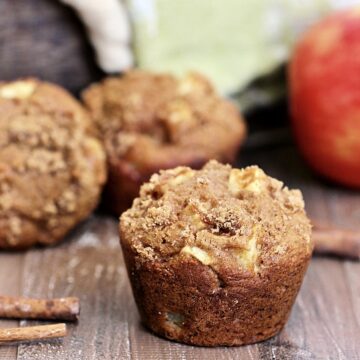 Single spiced apple muffin on a wooden board with more muffins and a red apple in the background.