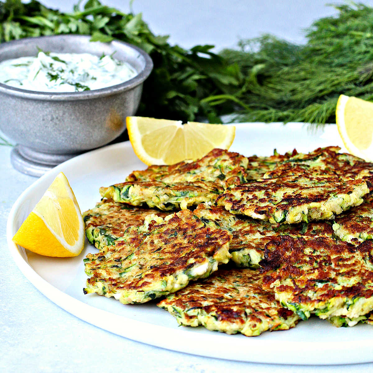 Greek zucchini fritters on a plate with lemon wedges. A bowl of tzatziki on the side.