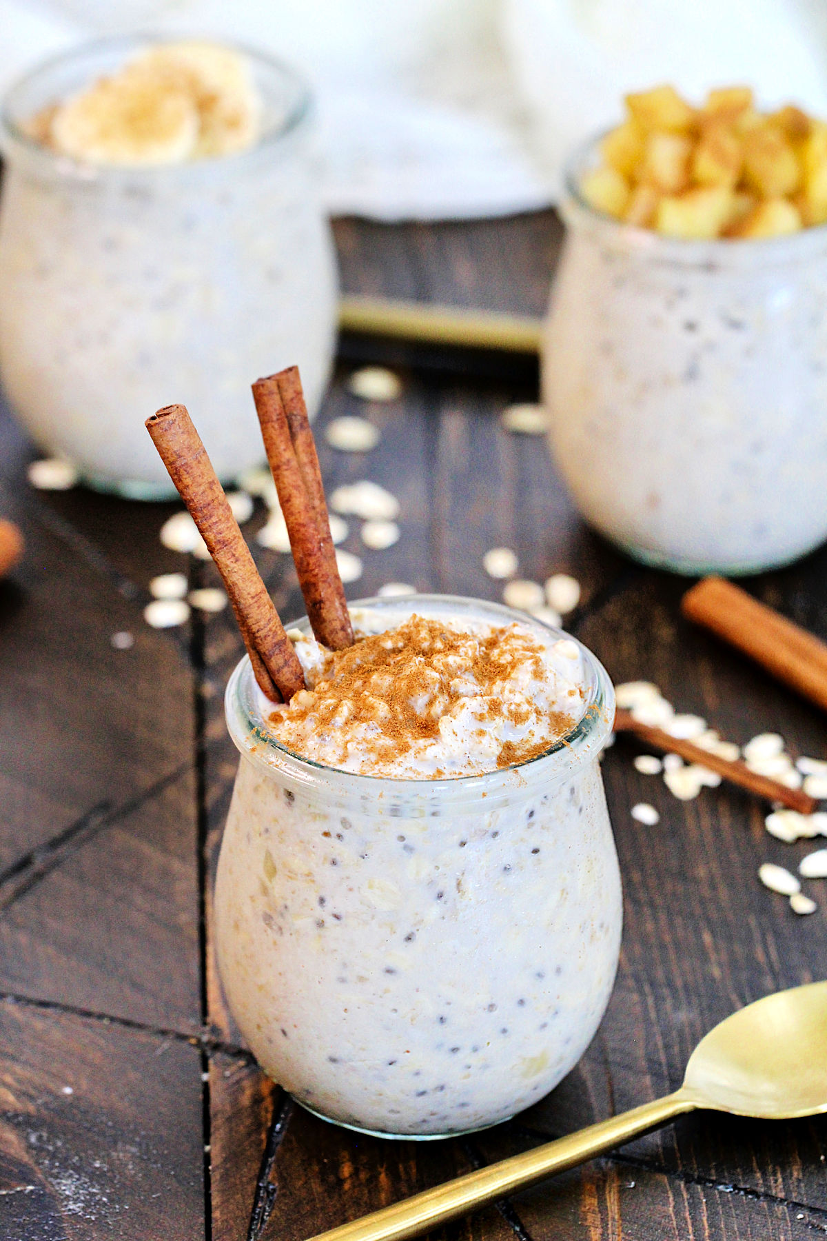 Cinnamon Overnight Oats in a glass jar topped with cinnamon sticks with a gold spoon and oats lying around it.