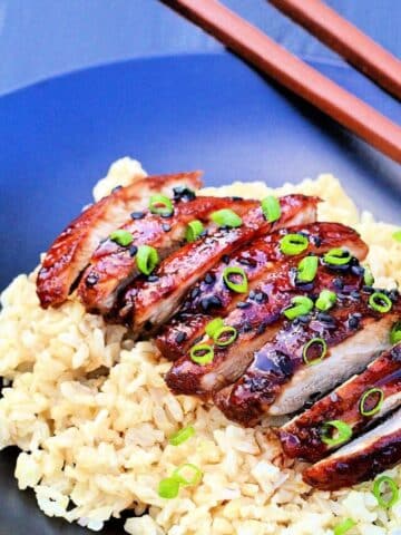 Sliced chicken teriyaki on a bed of brown rice with brown chopsticks nearby.