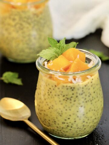 Two mango chia puddings topped with chunks of fresh mango in glass jars with a gold spoon and mint leaves around them.
