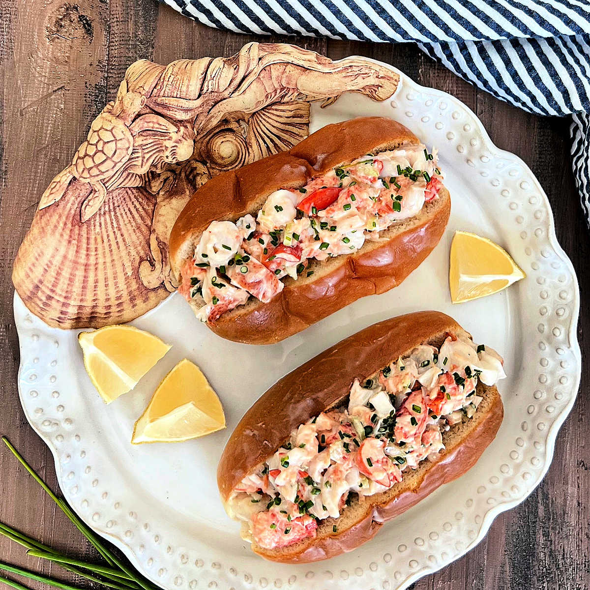 Two lobster rolls on a white plate with lemon wedges.
