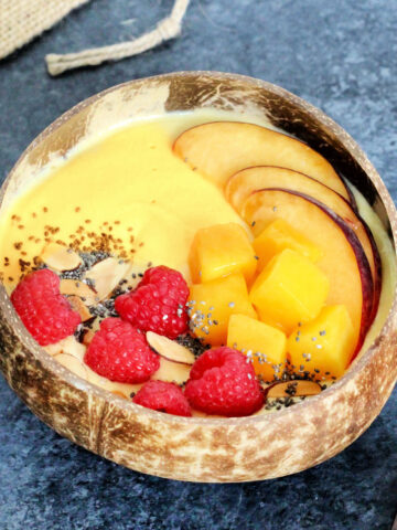 A Mango Smoothie Bowl in a wooden bowl surrounded by a wooden spoon and raspberries.