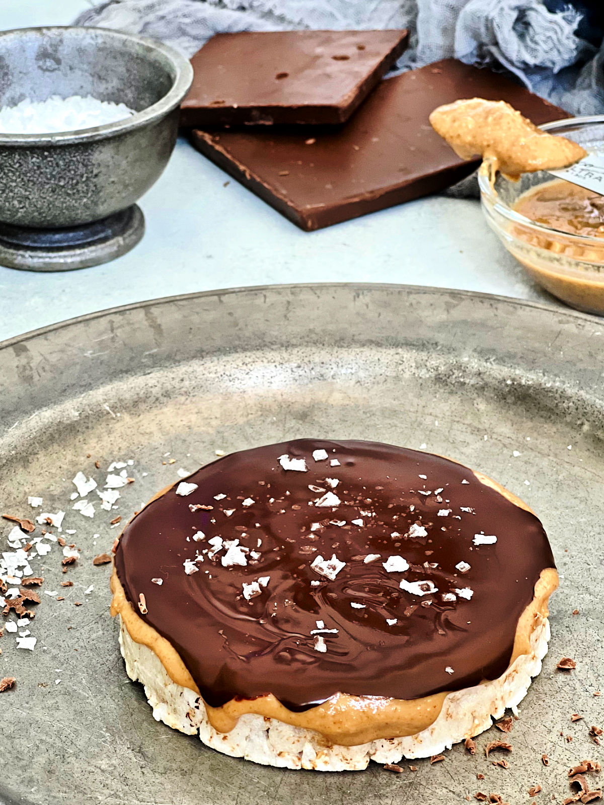 Giant peanut butter cup rice cake on a silver plate with chocolate, peanut butter and salt in the background.