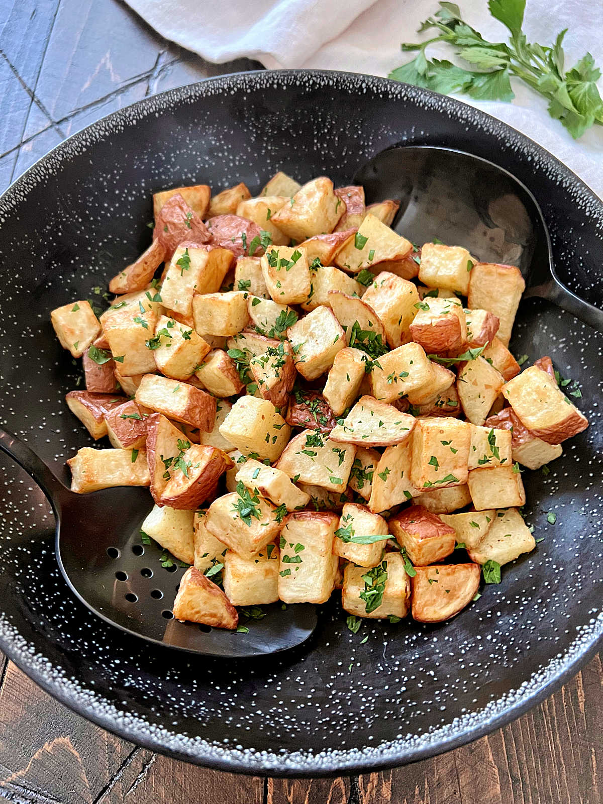 Air fryer diced potatoes garnished with parsley in a black bowl with two black spoons. 