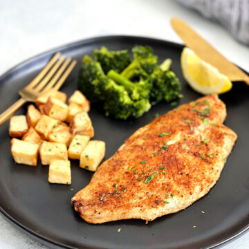 Air fryer tilapia on a black plate with diced potatoes, broccoli and a lemon wedge.
