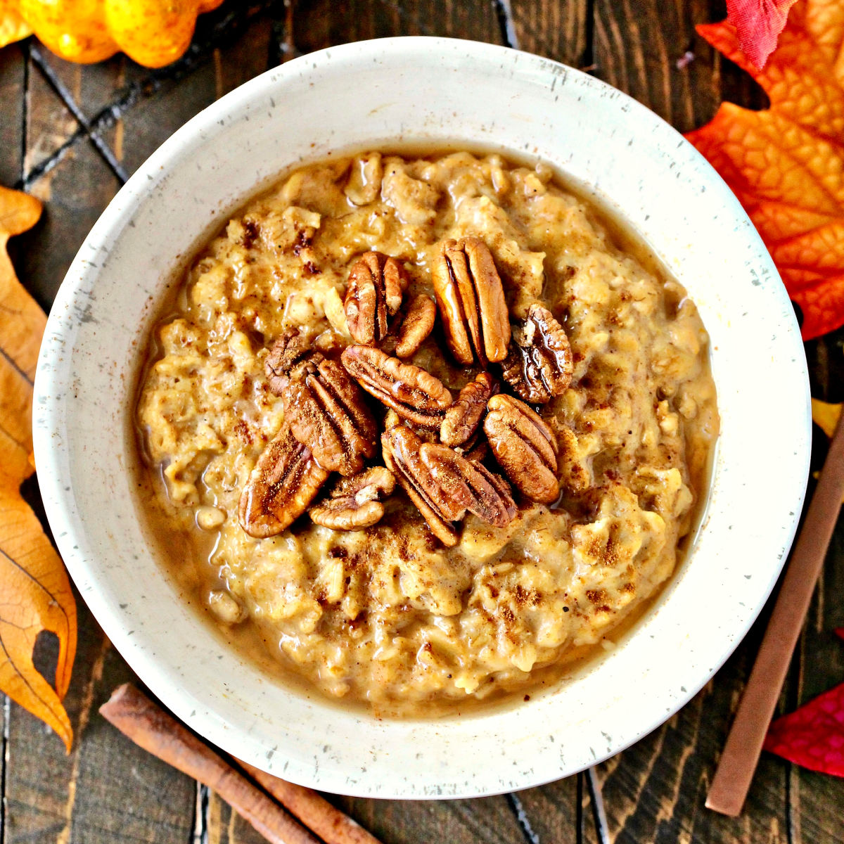Overhead shot of Pumpkin Spice Oatmeal in a white bowl on a brown wooden board with scattered fall leaves and gourds.