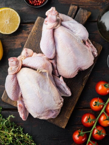 Uncooked whole chickens on wooden cutting board surrounded by fresh ingredients