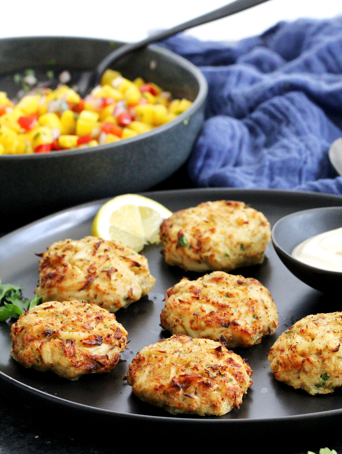 Maryland-style Old Bay crab cakes on a black plate with a bowl of mango salsa in the background.