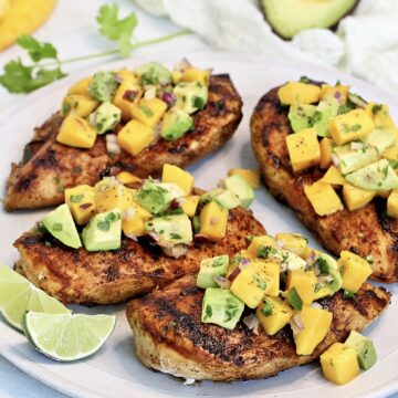 Grilled chicken with mango avocado salsa on a white plate with mango and avocado in the background.