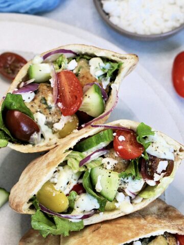 Three Greek pita sandwiches on a round plate with a bowl of feta cheese next to it.