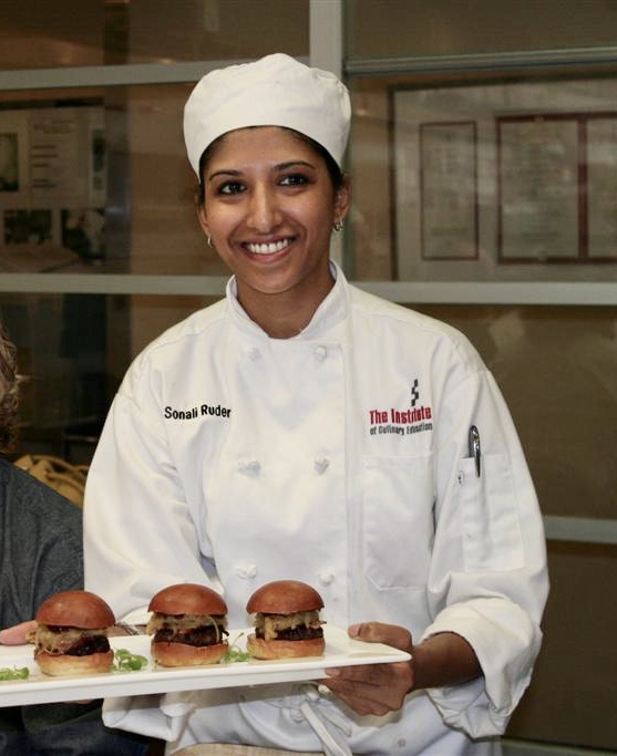 The Foodie Physician, Dr. Sonali Ruder in a chef's coat and hat holding a tray of hamburger sliders.