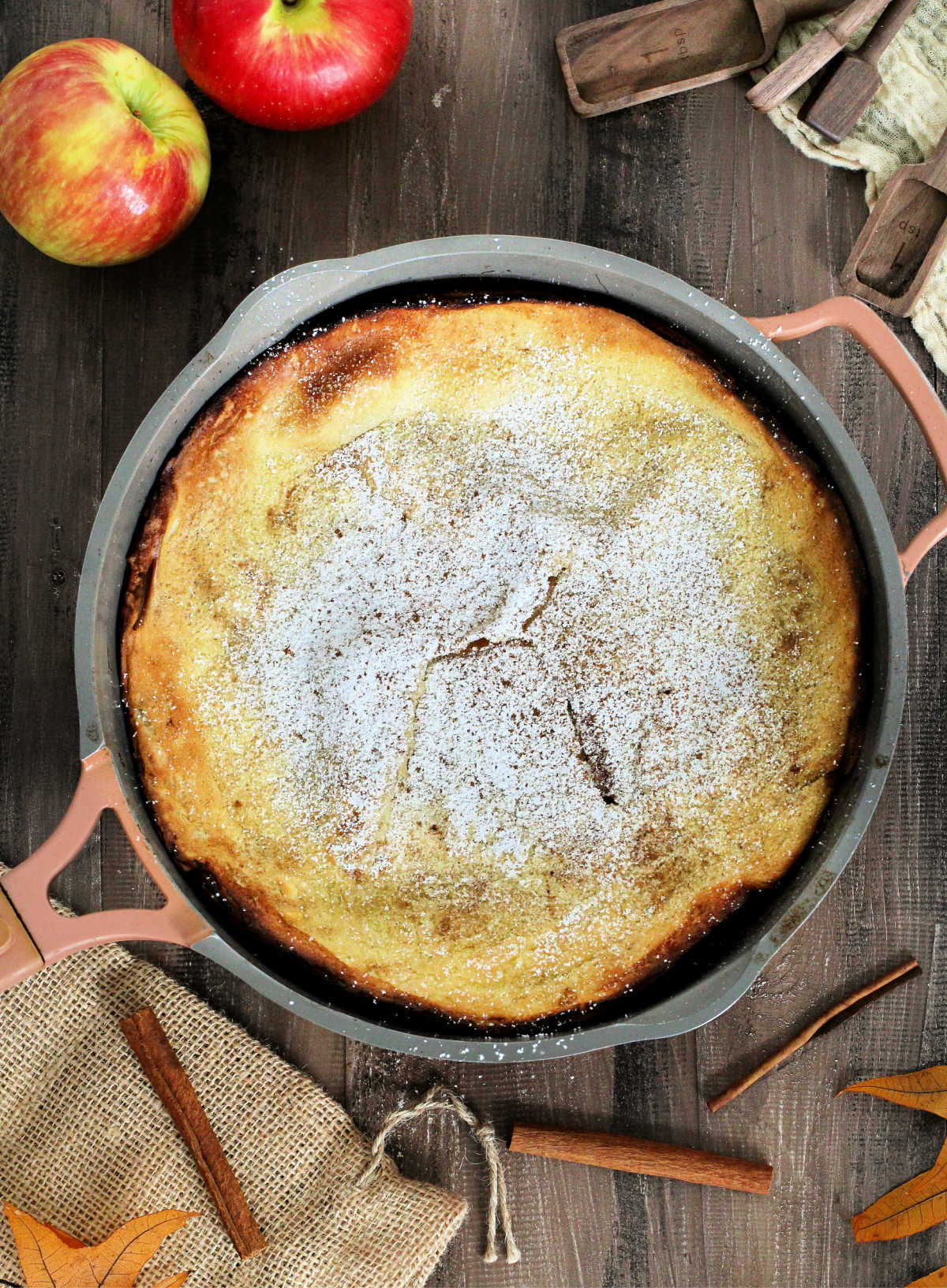 An Apple Dutch Baby in a pan on a wooden board surrounded by apples, cinnamon sticks and measuring spoons.