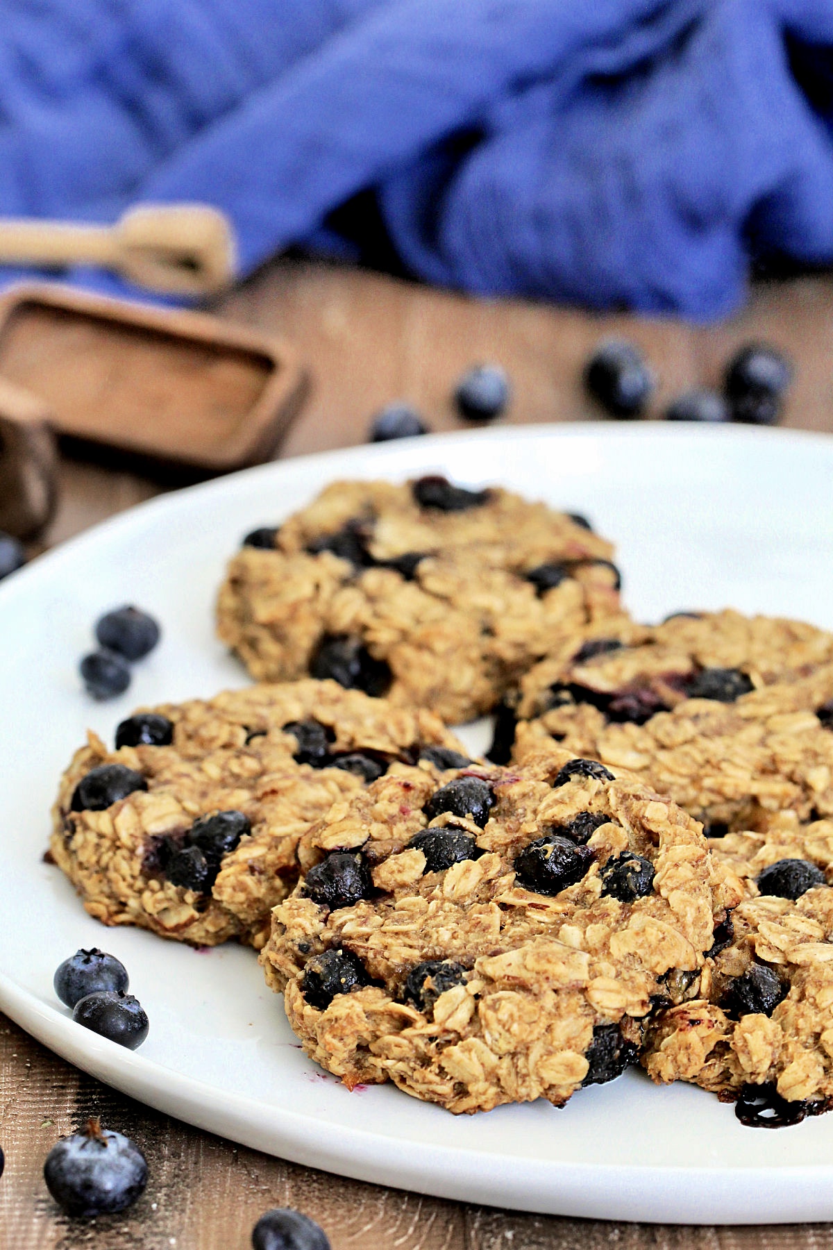 Blueberry oatmeal cookies on white plate surrounded by blueberries, wooden measuring spoons and a blue napkin.