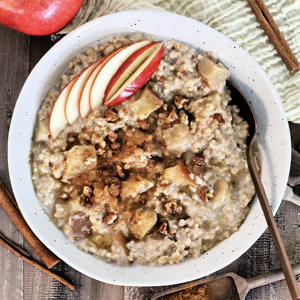 Easy Crockpot Apple Cinnamon Oatmeal in a bowl garnished with apple slices and cinnamon on a wooden board with an apple, cinnamon sticks and measuring spoons around it.