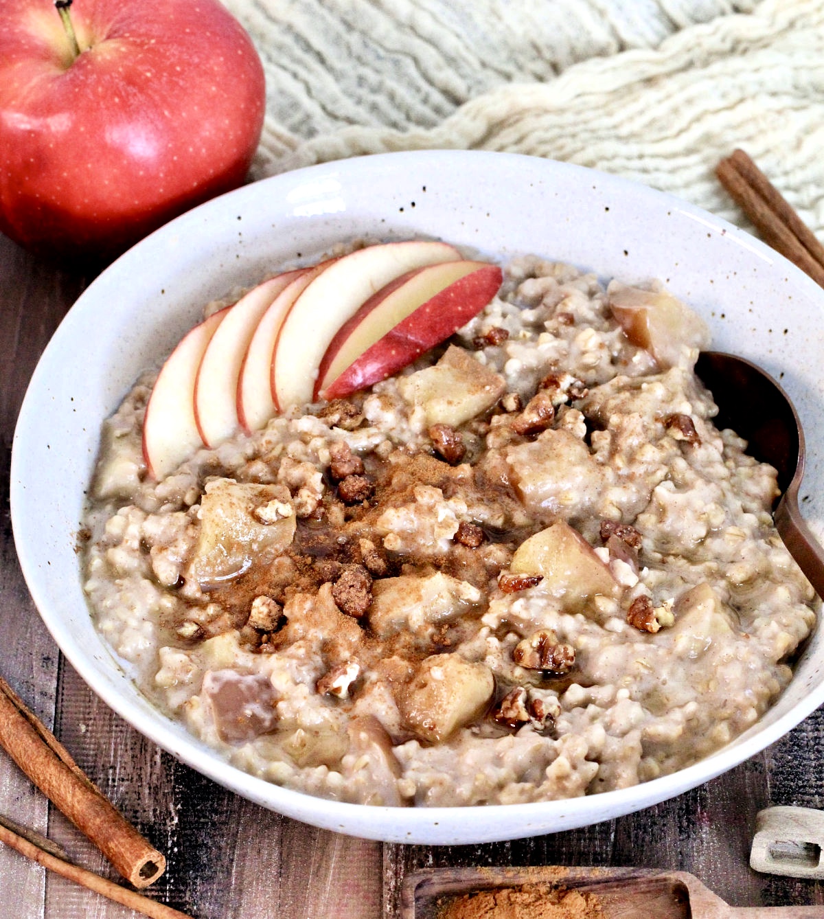 Easy Crockpot Apple Cinnamon Oatmeal in a bowl garnished with apple slices and cinnamon on a wooden board with an apple, cinnamon sticks and measuring spoons around it.