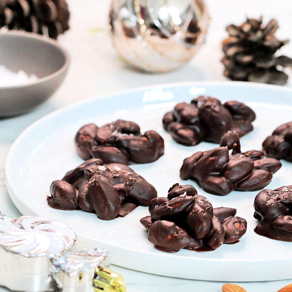 Chocolate almond clusters on a white plate with pinecones and silver ornaments around it.