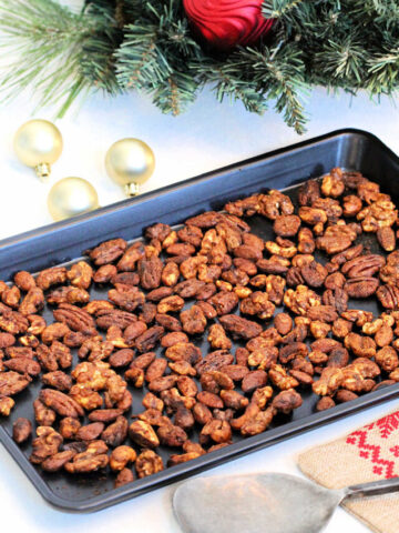 Holiday spiced nuts a black baking sheet in front of a wreath with ornaments around it.