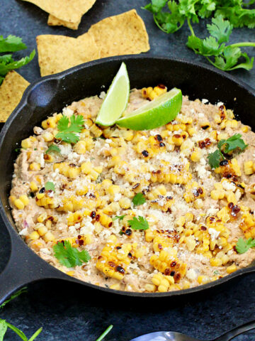 Mexican street corn dip in a cast iron skillet with tortilla chips, lime wedges and cilantro around it.