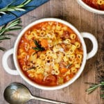 A bowl of pasta e fagioli soup on a wooden board surrounded by rosemary and a blue napkin.