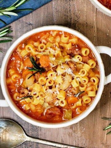 A bowl of pasta e fagioli soup on a wooden board surrounded by rosemary and a blue napkin.