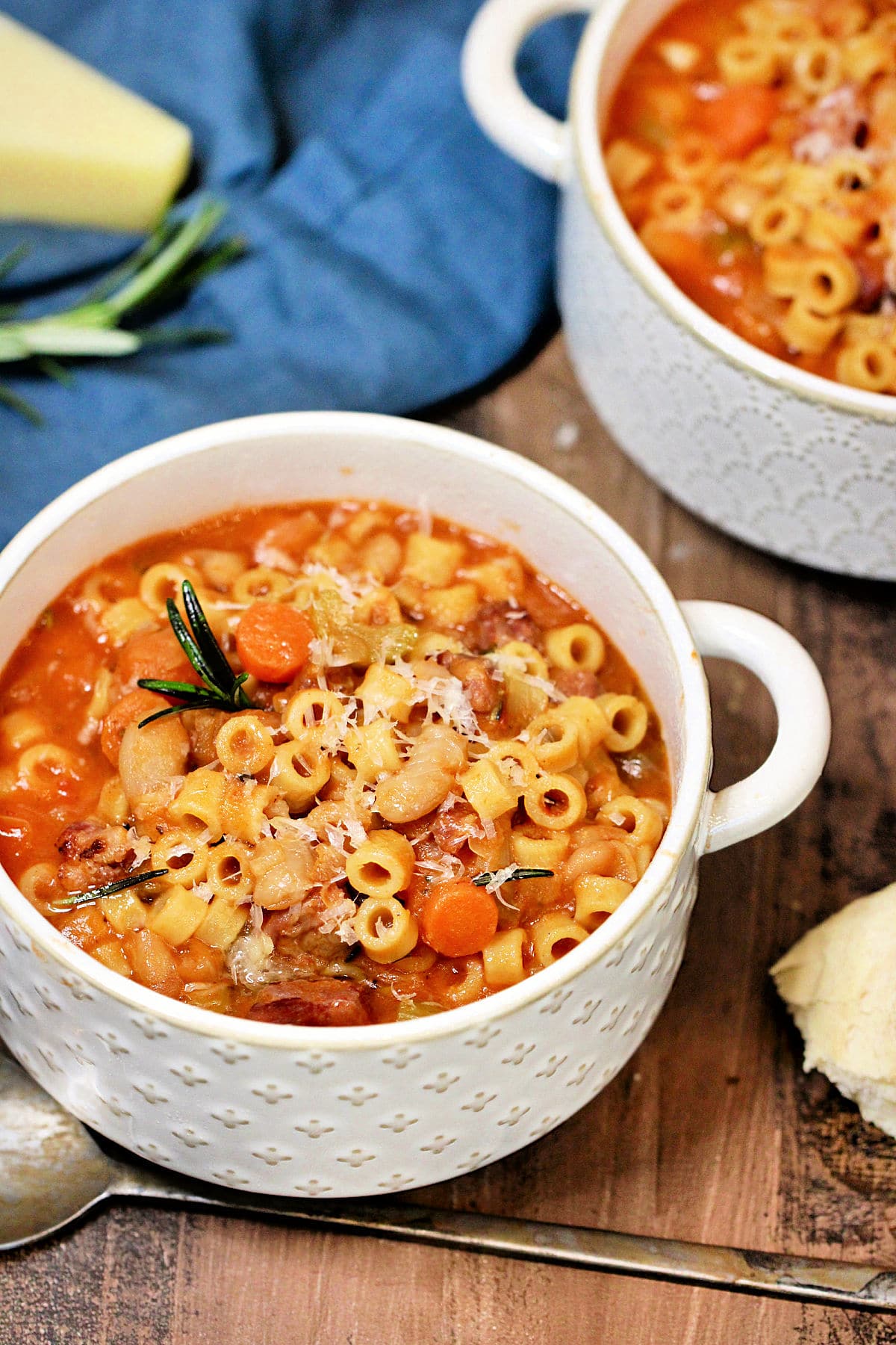 A bowl of pasta e fagioli soup on a wooden board surrounded by parmesan cheese, rosemary and a blue napkin.