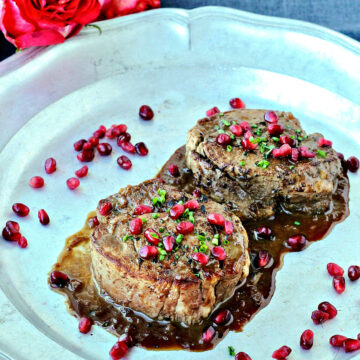Two filet mignons with pomegranate sauce on a silver platter with roses lying nearby.