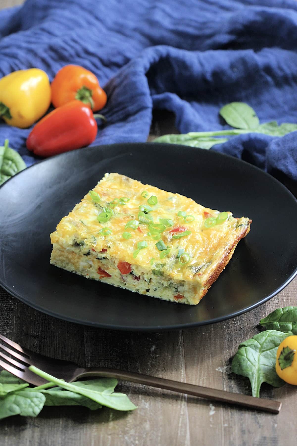 A slice of high protein cottage cheese egg bake on a black round dish surrounded by a blue napkin, colorful peppers and spinach leaves.